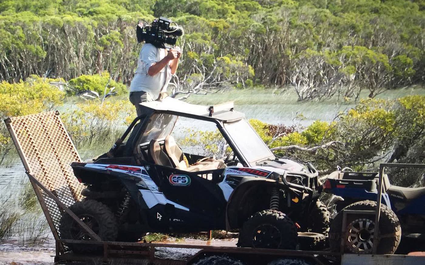A camera operator standing on a side-by-side as it crosses a flooded track