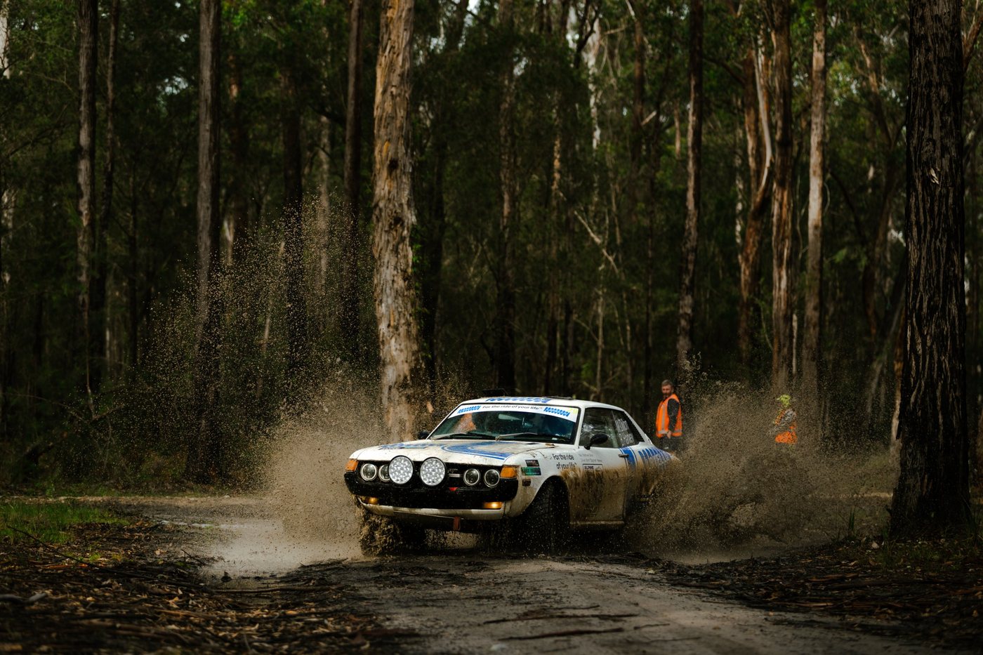 The Lovells rally car through a water splash on a forest stage, marshals watching